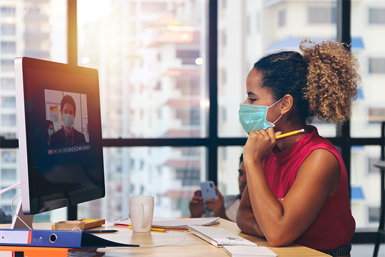 Woman sitting at desk with mask on looking at computer and video chatting with man also wearing a mask