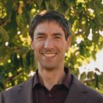 White appearing man with short brown hair with a blurred tree in the background