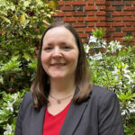 light-skinned appearing woman with brown hair, grey blazer over a red shirt, standing in front of an azalea bush with white blooms