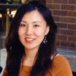 light-skinned woman with long black hair and an orange shirt with a brick wall background in half the photo