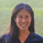 light brown skinned appearing woman with long black hair and a blue shirt on in front of a blurred green background