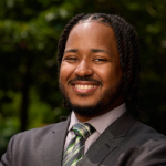 Dark skinned appearing male with black hair and a mustache with a suit and tie on in front of a dark green background