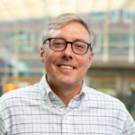 White appearing man with black-rimmed eyeglasses in front of a light, blurry background